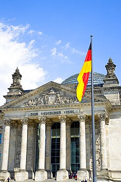 Germany, Berlin, Mitte, The Reichstag building in Tiergarten with the inscrption Dem Deucschen Volke, For the German People, on the facade above the columns at the entrance with a German flag on a flagpole and Norman Foster's glass roof dome behind.