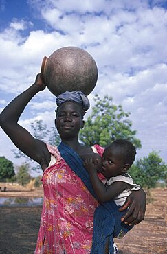 BURKINA FASO Children Woman breastfeeding child in sling at her side while carrying water pot on her head.