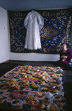 TAJIKISTAN  Wedding A white wedding dress hangs on the wall in front of a rug  and a large display of food is laid out awaiting guests from a village wedding