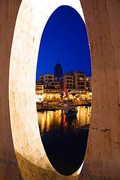 Saint Julians Bay waterfront at dusk with illuminated restaurants in front of apartments and the Hilton Tower office complex with fishing boats in the harbour in the foreground seen through a spherical opening, Saint Julians, Malta