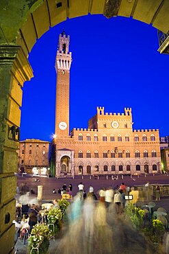 ITALY, Tuscany, Siena, The Torre del Mangia campanile belltower of the Palazzo Publico illuminated at night seen through the archway of the narrow sidestreet leading into the Piazza del Campo with people seated at tables and walking in the square