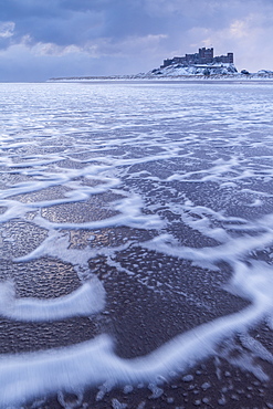 Foam patterns on Bamburgh Beach with a snow covered Bamburgh Castle on the horizon in winter, Bamburgh, Northumberland, England, United Kingdom, Europe