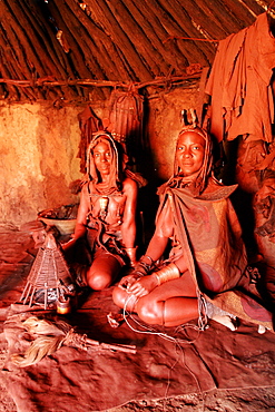 Two Himba women In goatskin clothes in their hut, working to keep the fire on the stone, Namibia, Africa