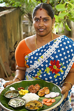 Mrs Vijayalakshmi Neelakandan with complete meal in a thali covered with a banana leaf with all the dishes - rasam, pickles, salad, seasoned yogurt, rice, meat stew, potatoes, salt, fried fish, curried eggs.