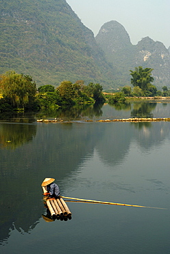 Chinese fisherman with a straw hat on a bamboo raft fishing near Yangshuo in the Yulong River in front of limestone cliffs, Yangshuo, Guilin, Guanxi, China