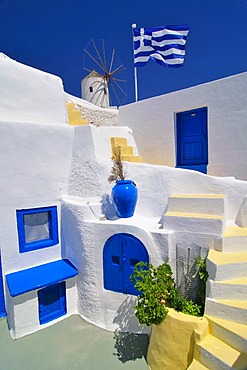 Blue and yellow inner courtyard with stairs, Greek flag blowing in the wind, typical Cycladic architecture, windmill behind, Oia, Ia, Santorini, Cyclades, Greece, Europe