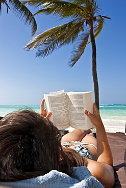 Young woman on a beach reading a book under palm trees