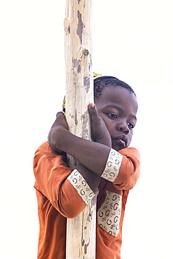 Portrait of a little boy wearing an orange abaya and a black and gold kofia embracing a wooden pole. In the late afternoon, children from a small village come to play by the sea, a white sand beach and turquoise sea on the east coast of Zanzibar, Tanzania.