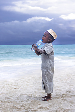 Little boy dressed in an abaya, light gray jdellaba and wearing a golden kofia, trying to drink water from a bottle. In the late afternoon, children from a small village come to play by the sea, a white sand beach and turquoise sea on the east coast of Zanzibar, Tanzania.