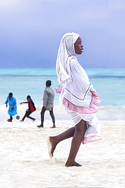 Young girl wearing white dress with pink ruffles and veil, white hijab. In the late afternoon, children from a small village come to play by the sea. White sand beach and turquoise sea on the east coast of Zanzibar, Tanzania.