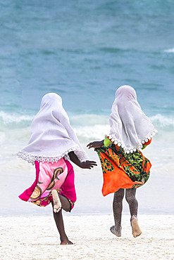 Two little girls dressed in colorful dresses and a white hijab run towards the sea. In the late afternoon, children from a small village come to play on the white sand beach and turquoise sea on the east coast of Zanzibar, Tanzania.