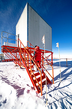 A technician goes into a raised square building on an orange metal structure. An Italian technician specialising in radio and telecommunications goes into the shelter that houses the VSAT antenna, the device that connects the station to the Internet by satellite. This is the station's only link with the outside world. Most of the buildings are elevated to prevent the formation of snowdrifts caused by wind-blown snow. Concordia Antarctic Research Station, Dome C plateau, East Antarctica.