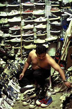 In cramped conditions at the Chatuchak Market, Bangkok, a Thai trader is searching through many pairs of 2nd hand trainers, some of which take pride of place covering the walls of his stall. Bangkok, Thailand